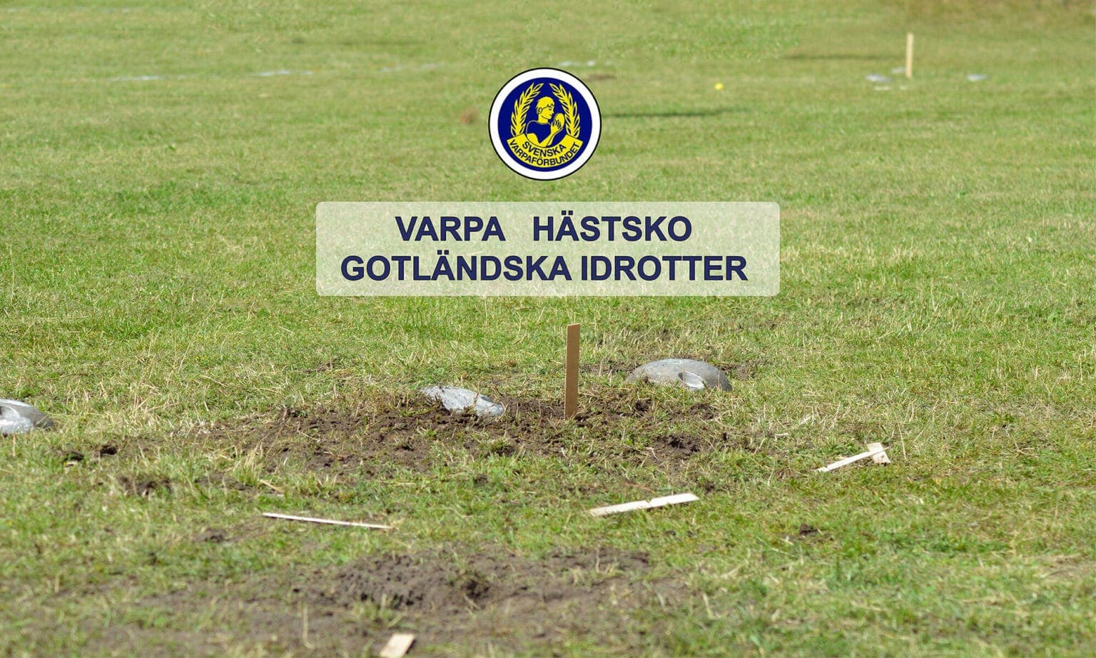 Varpabana med två stenar — en blank metallvarpa och en mörk gjuten sten — och en uppställd trästicka som mål, fotograferad på den gröna gräsplanen